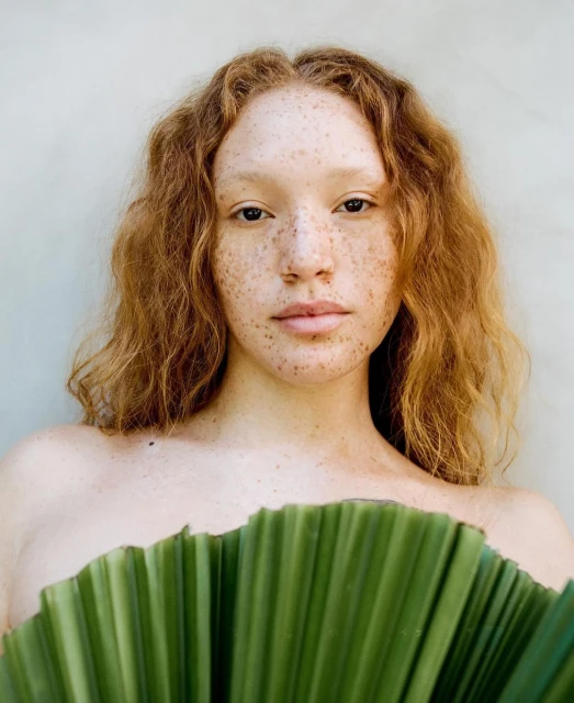 Portrait of a red-haired, freckled woman holding a large green leaf in front of her body against a neutral wall backdrop.