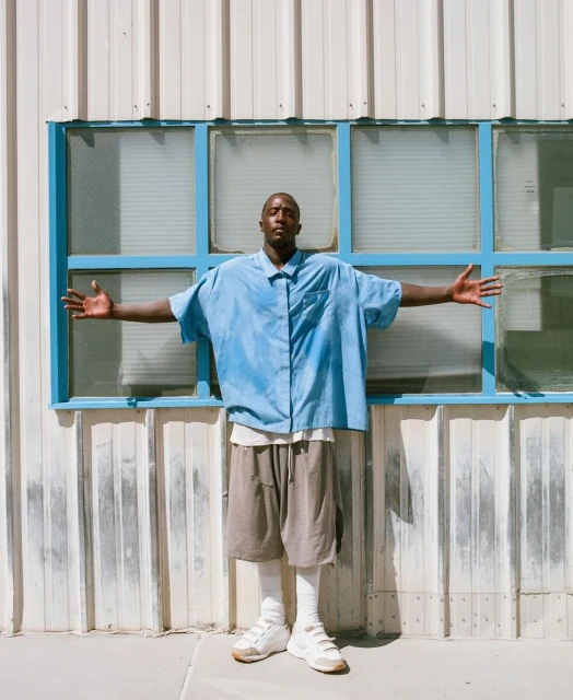 Man in oversized blue shirt and khaki shorts poses in front of weathered metal building with blue-framed windows, arms outstretched.