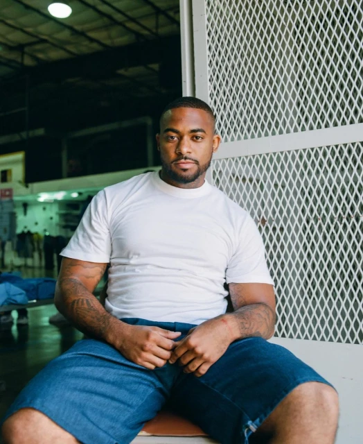 Portrait of a muscular, tatted African-American man in a white t-shirt and denim shorts, indoors with a metal grate background and natural light.