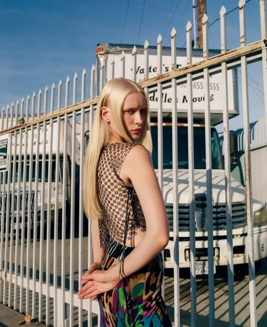 Fashion shot of a blonde adult female model in a patterned top and skirt, posing outdoors in front of a chain link fence and moving truck.