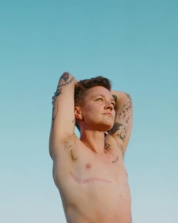 Person with short hair and chest scar posing against a blue sky backdrop, with tattoos visible on their arms and shoulders.
