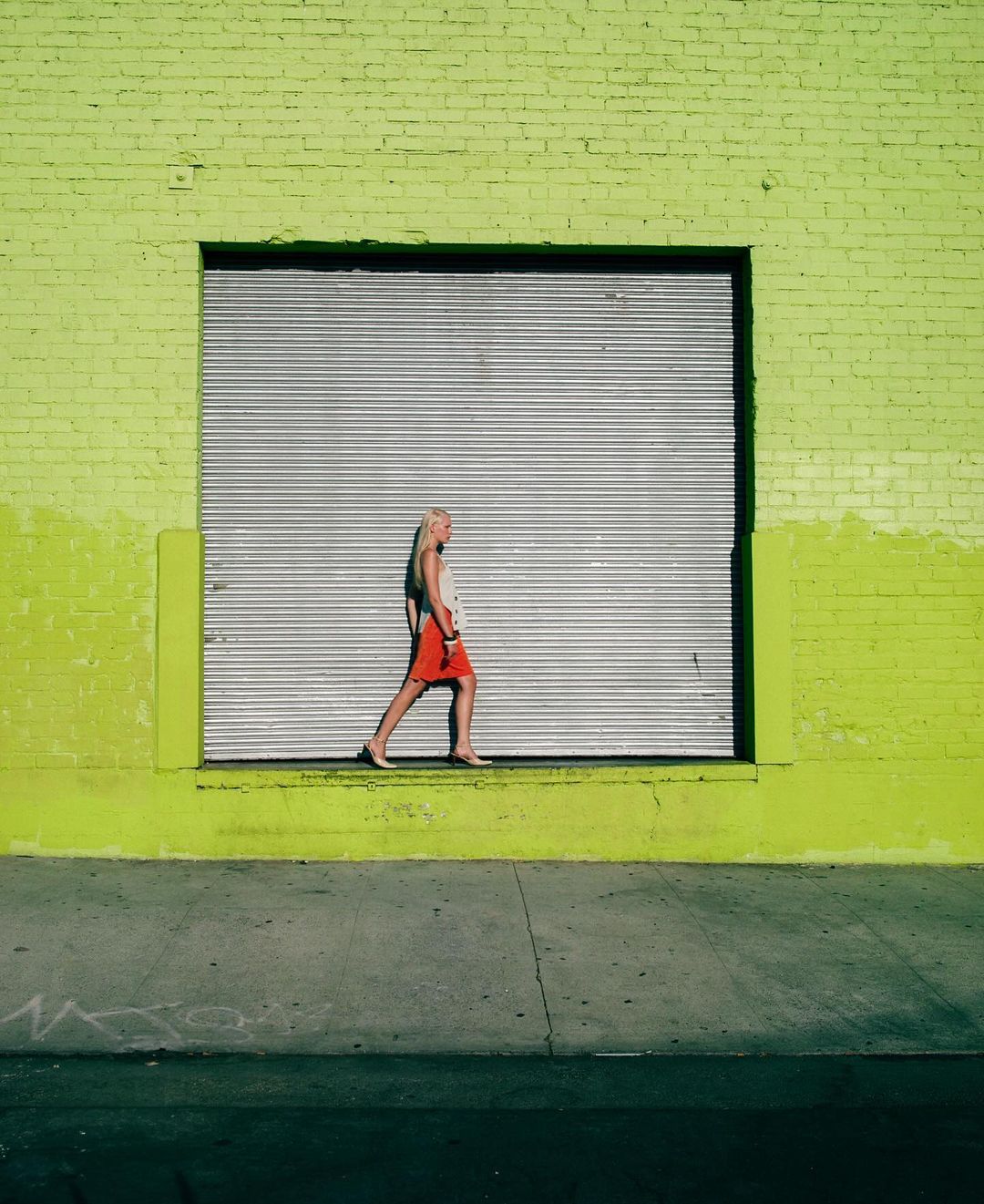 Young woman walking in front of a building with a closed silver corrugated steel door and bright green painted brick facade, urban setting.