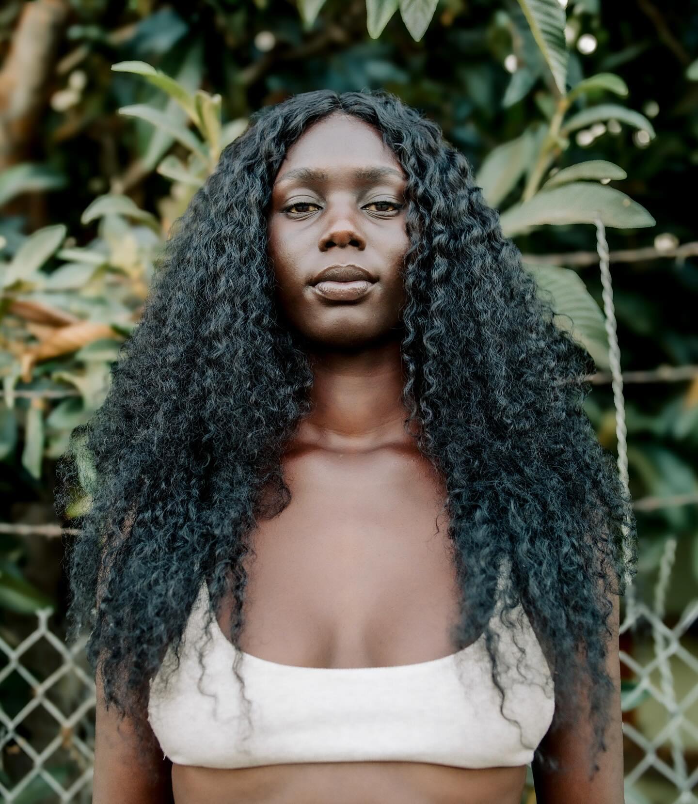 Close-up of a woman with dark skin and curly hair wearing a beige top, posing outdoors against a green foliage background.