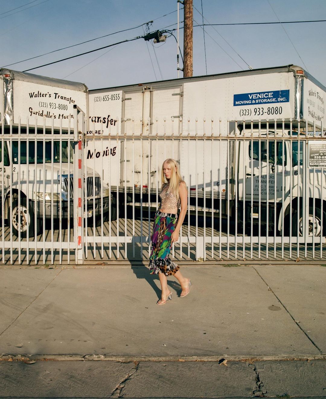 Woman in a colorful skirt and metallic top stands in front of a white metal gate and moving trucks on a city street.