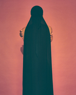 Back view of an adult female in a long, dark hooded dress against a coral pink background, arms crossed.