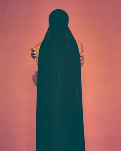 Back view of an adult female in a long, dark hooded dress against a coral pink background, arms crossed.