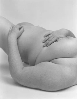 Nude adult female figure lies on a light surface, arms folded over her body, in a studio shot with soft lighting and monochromatic tones.