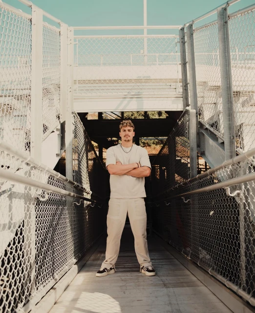 Full body portrait of a young adult male standing on a pedestrian bridge with his arms crossed and wearing casual clothing and sneakers.