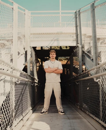 Full body portrait of a young adult male standing on a pedestrian bridge with his arms crossed and wearing casual clothing and sneakers.