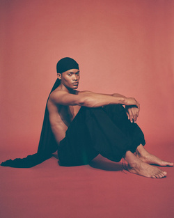 Portrait of a shirtless adult male in black pants and a durag, seated against a plain coral background in a studio.