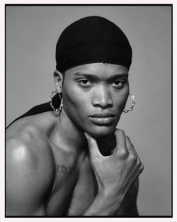 Striking black and white portrait of a shirtless person with a durag, hoop earrings, and hand on chin, set against a neutral gray background.