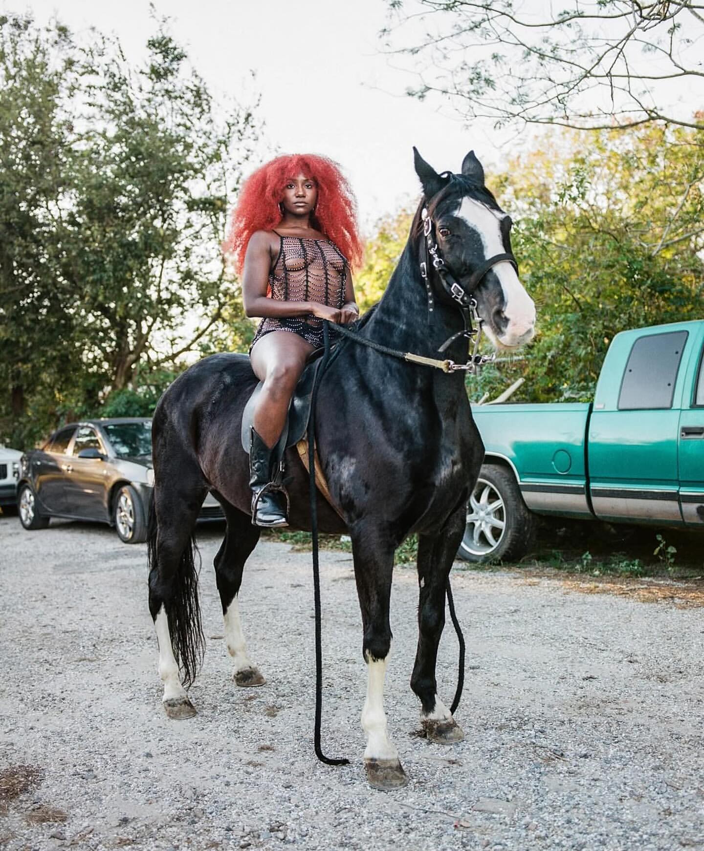 A black horse stands outdoors with a woman with red hair riding it wearing a dark, crocheted bathing suit and boots near vehicles.