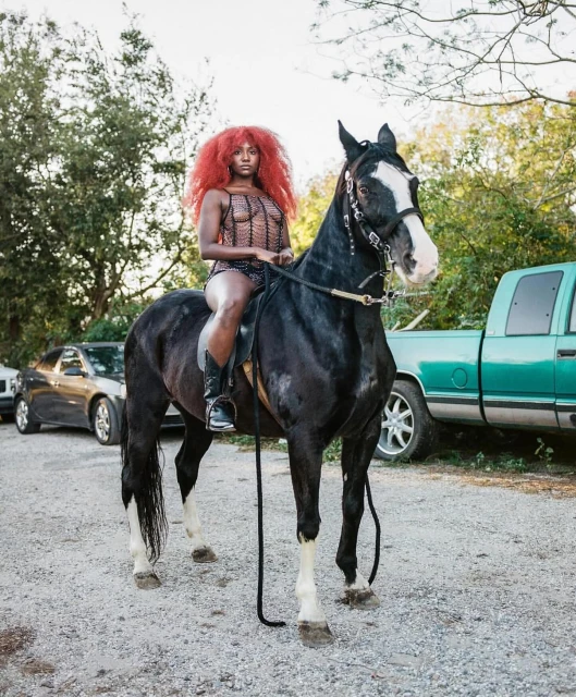A black horse stands outdoors with a woman with red hair riding it wearing a dark, crocheted bathing suit and boots near vehicles.