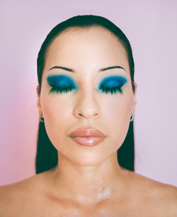 Headshot of a woman with eyes closed wearing vibrant blue eyeshadow, glossy lips, and neutral top against a light pink background.