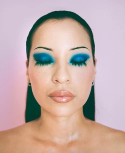 Headshot of a woman with eyes closed wearing vibrant blue eyeshadow, glossy lips, and neutral top against a light pink background.