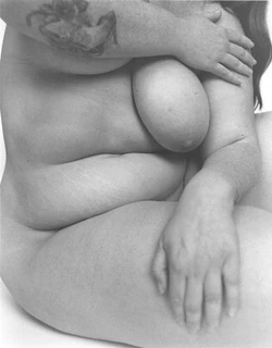 Nude female figure sitting with arms crossed in a black and white photograph showcasing curves and body positivity with skin texture.