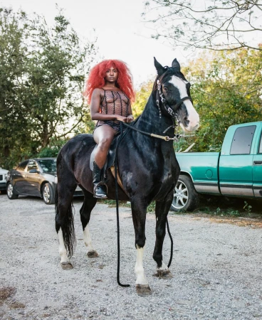 Black woman with vibrant red hair riding a dark horse outdoors with parked cars and trees in background