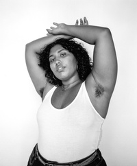 Black and white studio portrait of an adult woman with arm hair, wearing a white tank top and black pants, with neutral facial expression.