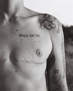 Monochromatic photograph of a bare-chested person with visible tattoos, scars, and writing on their torso and upper arms, with blurred background.
