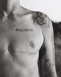 Monochromatic photograph of a bare-chested person with visible tattoos, scars, and writing on their torso and upper arms, with blurred background.