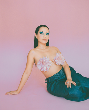 Woman with blue eyeshadow and pink flower pasties wearing dark trousers against a pink gradient background.