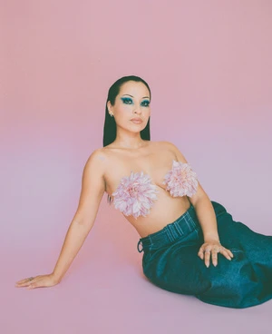 Woman with blue eyeshadow and pink flower pasties wearing dark trousers against a pink gradient background.