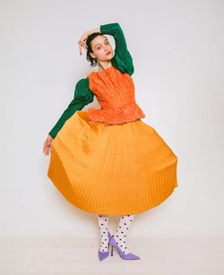 A fashionably dressed young woman in a textured orange top and matching skirt poses in a photo studio against a plain white backdrop.