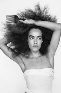 Portrait of a woman with large afro hairstyle holding a hair pick and wearing a strapless top against a white background in studio lighting.