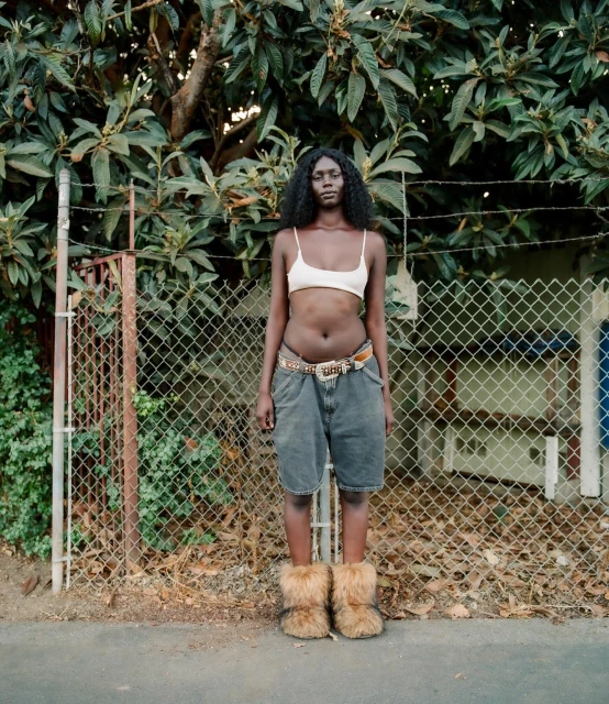 A slender Black female model stands in front of a chainlink fence wearing denim shorts, a top, and furry boots, outdoors in daytime.