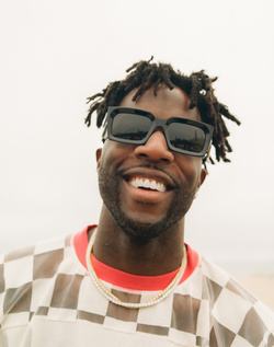Portrait of a smiling Black man with dreadlocks wearing chunky black sunglasses and a patterned shirt, shot by Maiwenn Raoult.