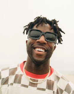 Portrait of a smiling Black man with dreadlocks wearing chunky black sunglasses and a patterned shirt, shot by Maiwenn Raoult.