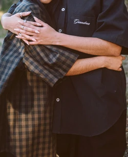 Close-up of two people embracing warmly, one wearing a brown plaid shirt and the other in a black button-down shirt with stitched text.