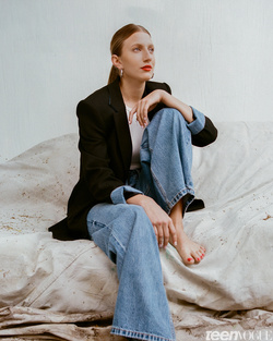 Woman sitting barefoot on a cushioned surface in a black blazer and wide-leg jeans. Her red lipstick stands out in the medium shot.