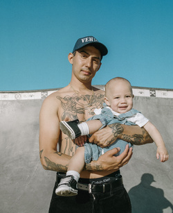 Shirtless man in baseball cap holds toddler wearing denim overalls in an outdoor skatepark under blue sky. Tattoos on man's chest and arms visible.