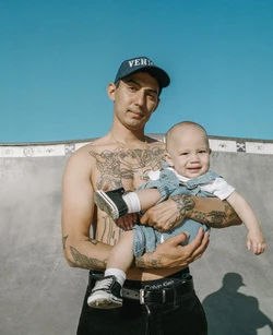 Shirtless man in baseball cap holds toddler wearing denim overalls in an outdoor skatepark under blue sky. Tattoos on man's chest and arms visible.