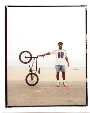 Man in a t-shirt and shorts stands on a beach holding a BMX bike in one hand, with ocean and sky in the background on a sunny day.