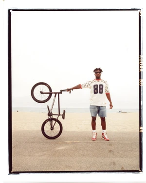 Man in a t-shirt and shorts stands on a beach holding a BMX bike in one hand, with ocean and sky in the background on a sunny day.