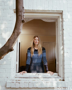 Woman stands in a window in a blue pattern dress. White brick, tree branch, interior room are visible. Natural light, medium shot.