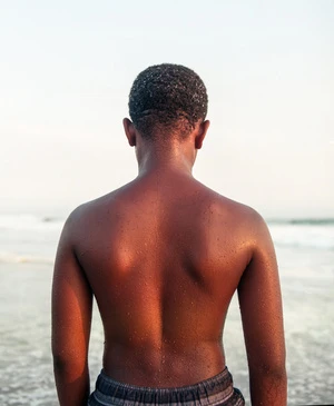 Rear view of a young Black man with water droplets on his back standing outdoors near the ocean by Maiwenn Raoult