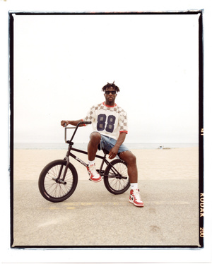 African American man sits on a BMX bike on a concrete surface with an ocean view, wearing shorts, sneakers, and a checkerboard shirt.