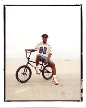 African American man sits on a BMX bike on a concrete surface with an ocean view, wearing shorts, sneakers, and a checkerboard shirt.