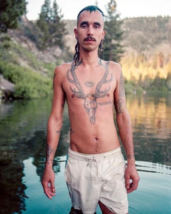 Young adult male standing in a mountain lake with tattoos visible on his chest, arms, and neck, with trees and mountains in the background.