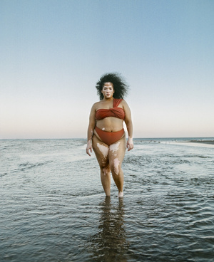 A woman with vitiligo in a red-brown swimsuit stands in the ocean under a blue sky, facing the camera with a neutral expression.