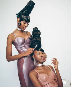 Two African American women pose in shiny lilac dresses with elaborate black hairstyles against a white backdrop in a studio shot.