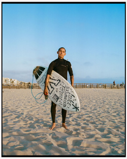 Surfer wearing a black wetsuit holding a patterned surfboard on a sandy beach under a clear sky in Los Angeles by Maiwenn Raoult