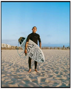 Surfer wearing a black wetsuit holding a patterned surfboard on a sandy beach under a clear sky in Los Angeles by Maiwenn Raoult