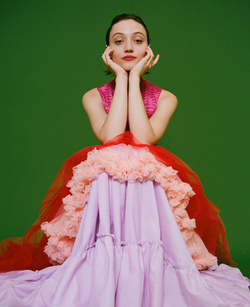 Portrait of a young woman in a colorful dress posing against a green background. The model rests her chin in her hands.