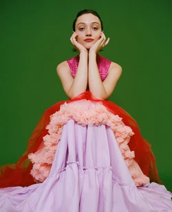 Portrait of a young woman in a colorful dress posing against a green background. The model rests her chin in her hands.