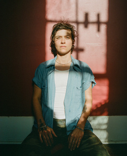 Adult woman with short brown hair poses indoors in casual clothes in sunlight with shadows cast across her body and a faded red wall behind.