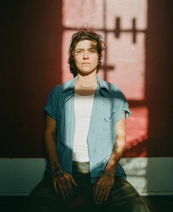 Adult woman with short brown hair poses indoors in casual clothes in sunlight with shadows cast across her body and a faded red wall behind.